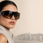 Woman wearing oversized black sunglasses and a beige sleeveless jacket against a beige backdrop with a faint world map overlay and Hong Kong skyline in the distance.