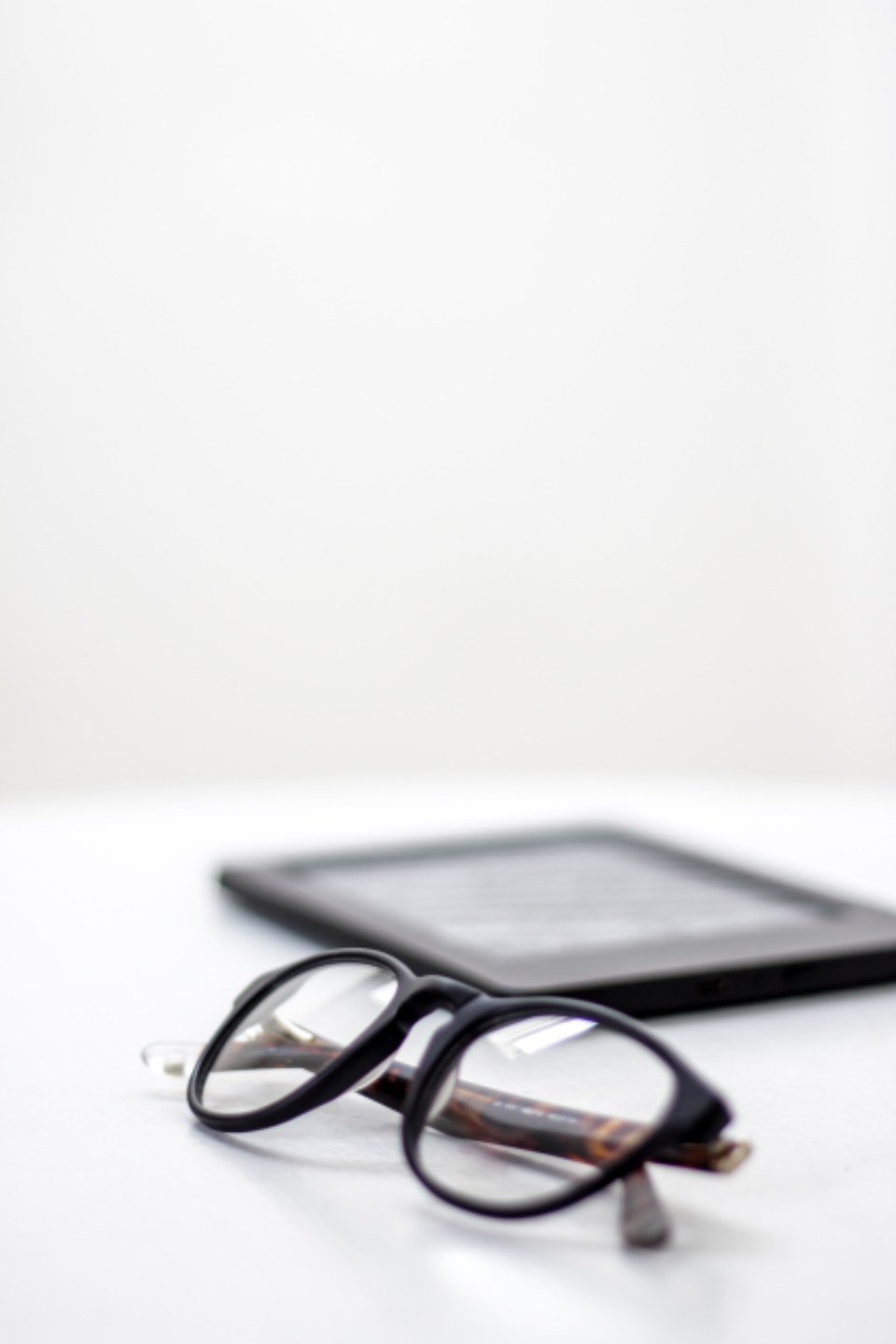 Black-rimmed glasses in the foreground on a white surface with a tablet blurred in the background.