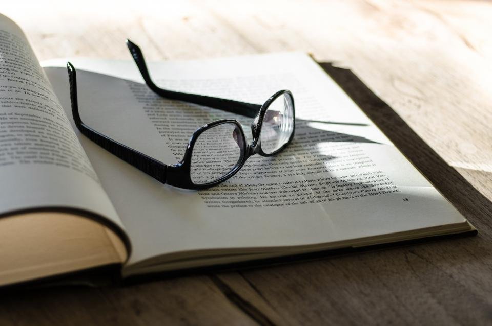 Glasses resting on open book pages on a wooden table in sunlight.