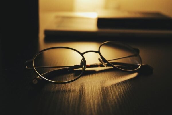 Reading glasses lying on a dark wooden desk with warm backlighting from a lamp behind them, a calm, contemplative scene.