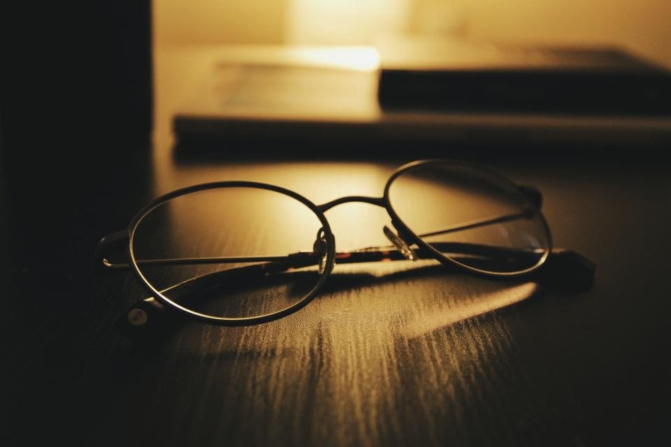 Reading glasses lying on a dark wooden desk with warm backlighting from a lamp behind them, a calm, contemplative scene.