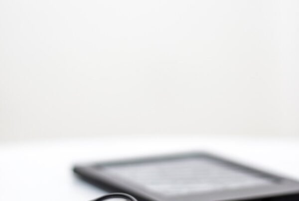 Eyeglasses with black frames resting on a white desk, with a blurred tablet in the background.