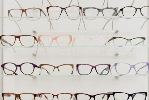 Eyewear replenishment cadence: unbranded optical frames on glass inventory shelves ready for reorder staging, no brand marks