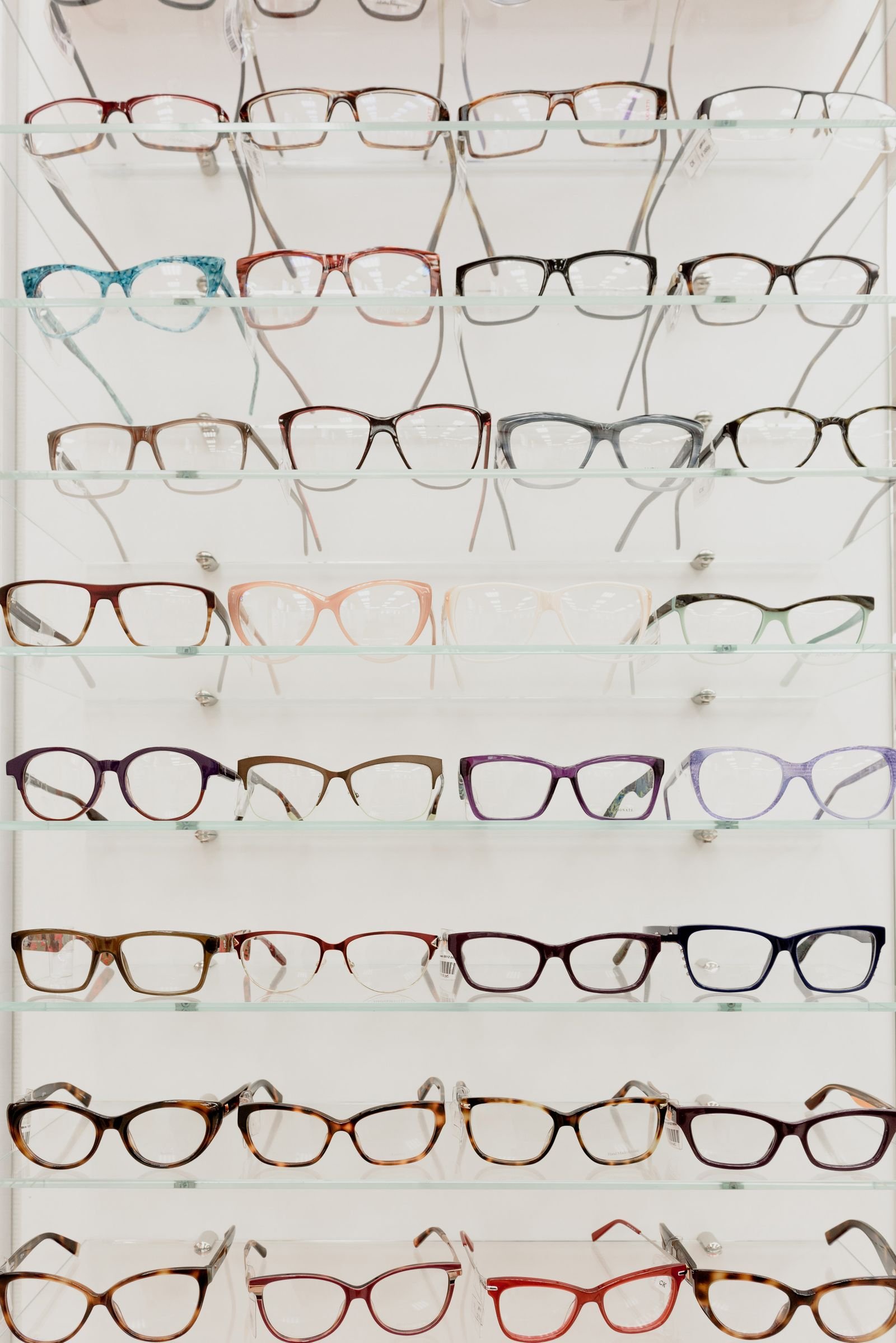Eyewear replenishment cadence: unbranded optical frames on glass inventory shelves ready for reorder staging, no brand marks