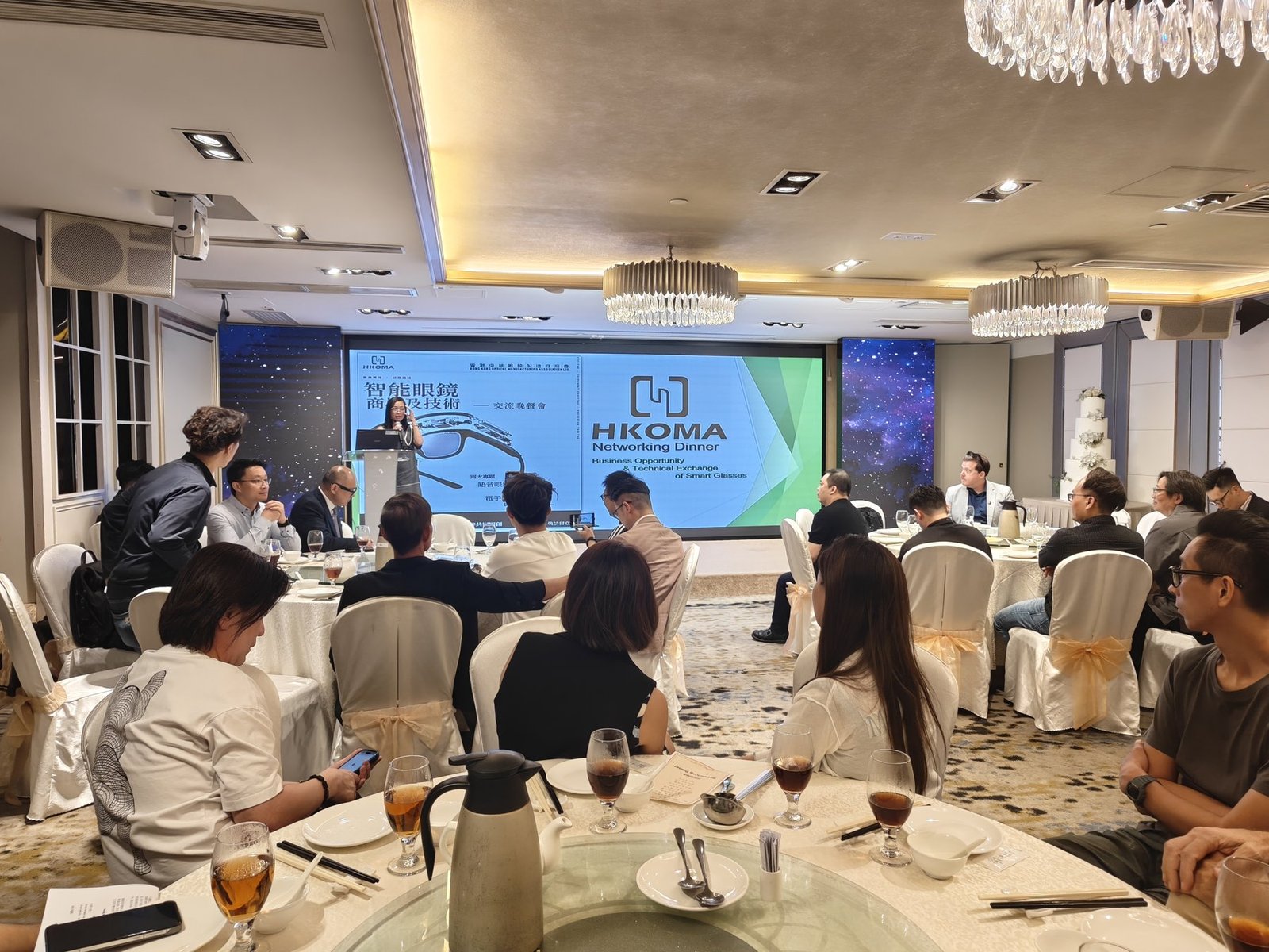 Networking dinner in a banquet hall; presenter at podium with large HKOMA screen, guests seated at round tables.