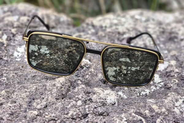 Gold-trimmed rectangular sunglasses with dark lenses resting on a rocky surface.