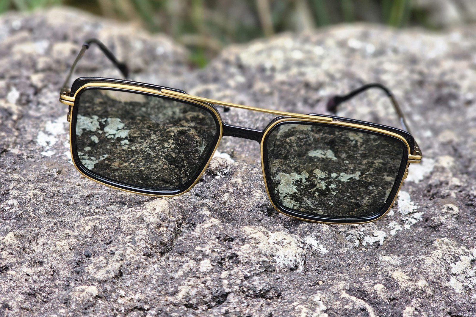 Gold-trimmed rectangular sunglasses with dark lenses resting on a rocky surface.